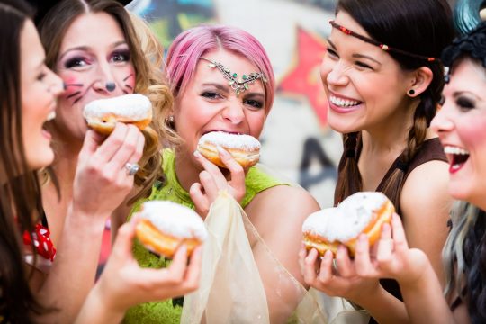 Group of women dressed up and smiling as they eat doughnuts at a Mardi Gras celebration