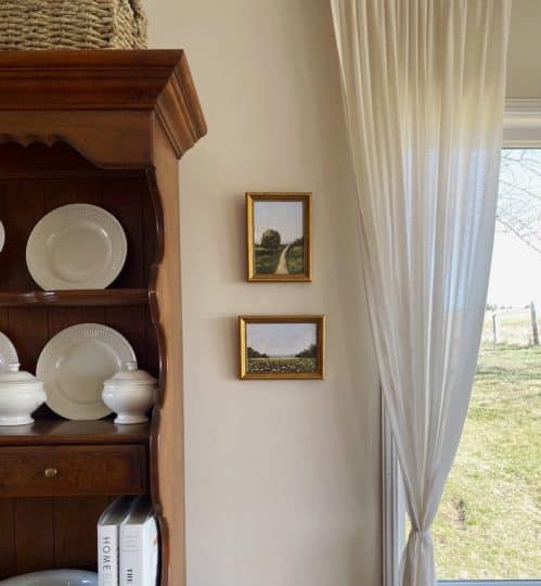 Two handmade pictures in gold frames hanging next to a wooden hutch