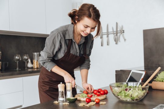 Image of young pretty woman standing in kitchen using tablet computer and cooking with the tomatoes and avocado.
